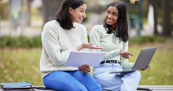 Two women sit on a bench sharing a computer and one of them is pointing at the screen.