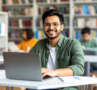 A smiling man with glasses working on a laptop in a library.