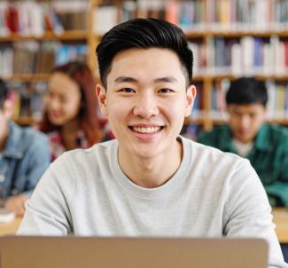 A young man smiling at the library on his laptop.