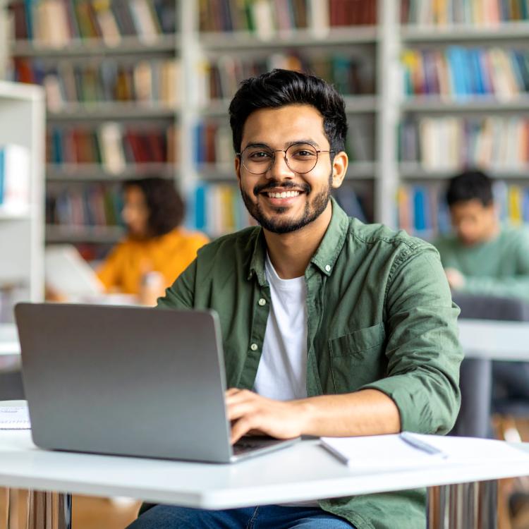 A smiling man with glasses working on a laptop in a library.