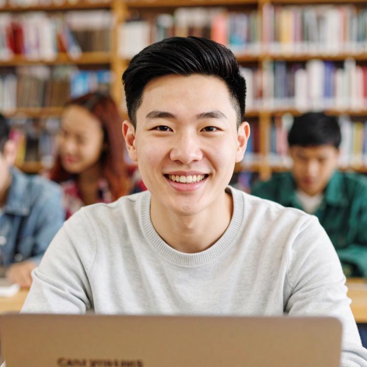 A young man smiling at the library on his laptop.